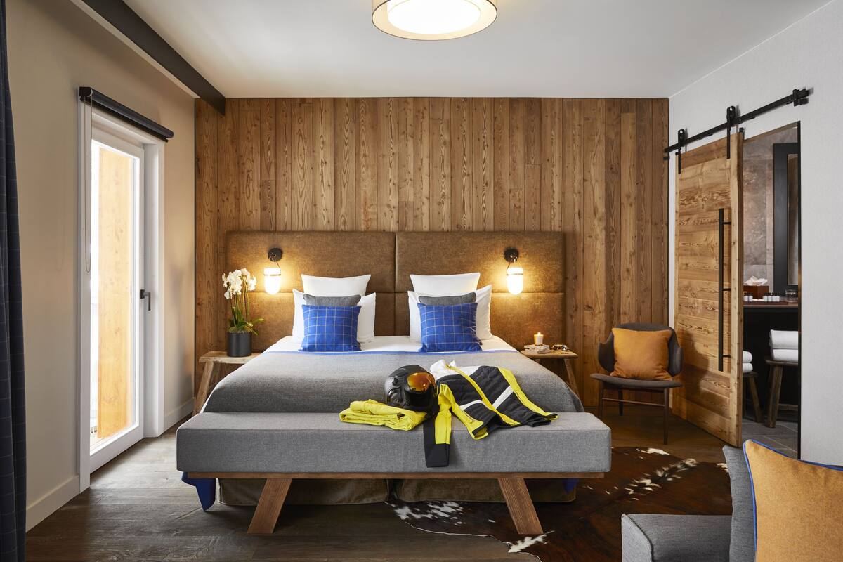 Una camera d'albergo moderna con parete in legno, un grande letto con materasso grigio e cuscini blu, un comodino con una lampada e una sedia.