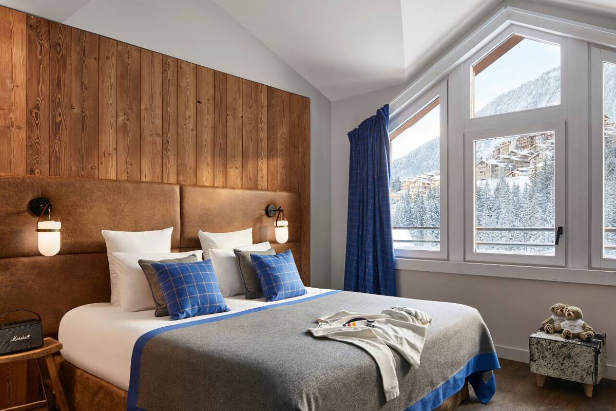 Una camera da letto con una testiera in legno, un letto grande con biancheria azzurra e bianca, un comodino con una lampada e una finestra che si affaccia su alberi innevati.