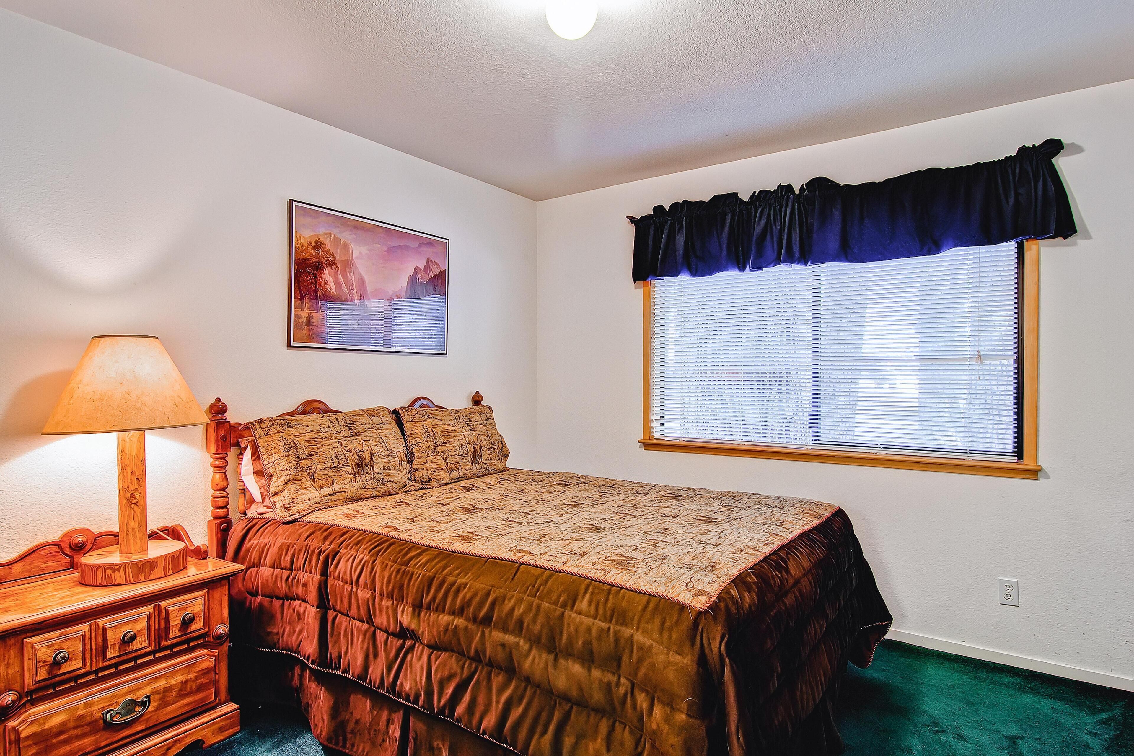 A bedroom with a bed, a nightstand, a lamp, a window with blinds, and a wall-mounted picture.