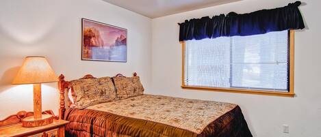 A bedroom with a bed, a nightstand, a lamp, a window with blinds, and a wall-mounted picture.