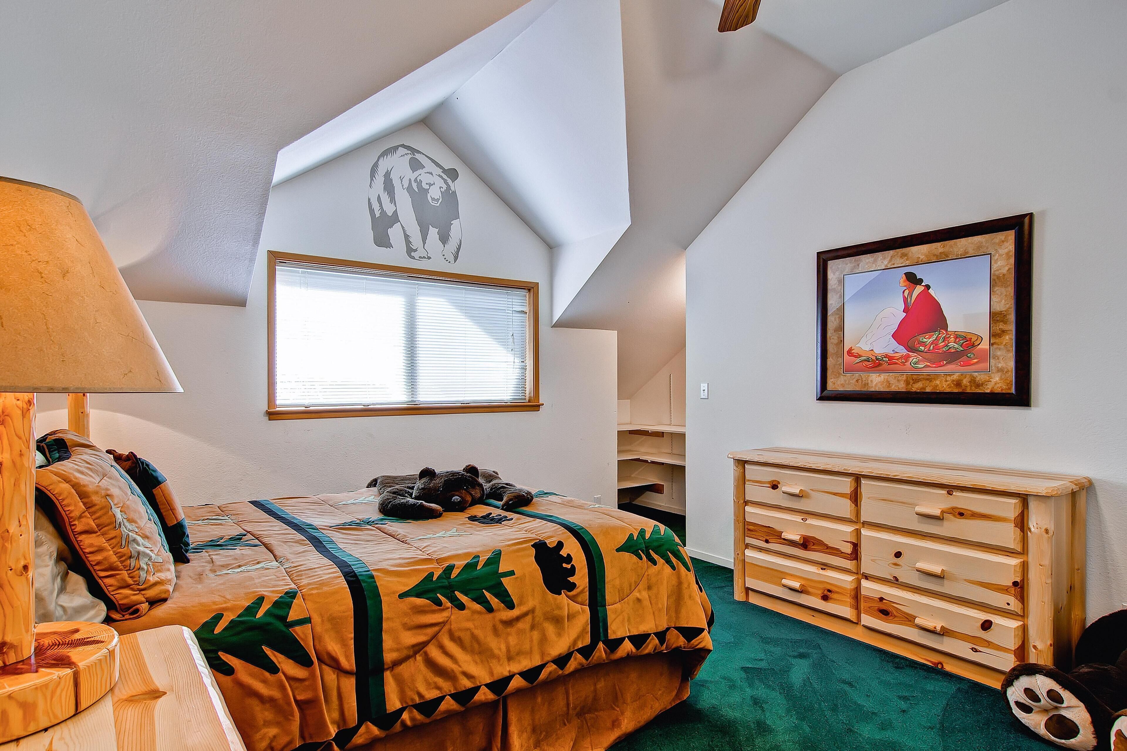 A bedroom with a sloped ceiling, a bed with a forest-themed bedspread, a wooden dresser, a window with blinds, and a bear mural on the wall.