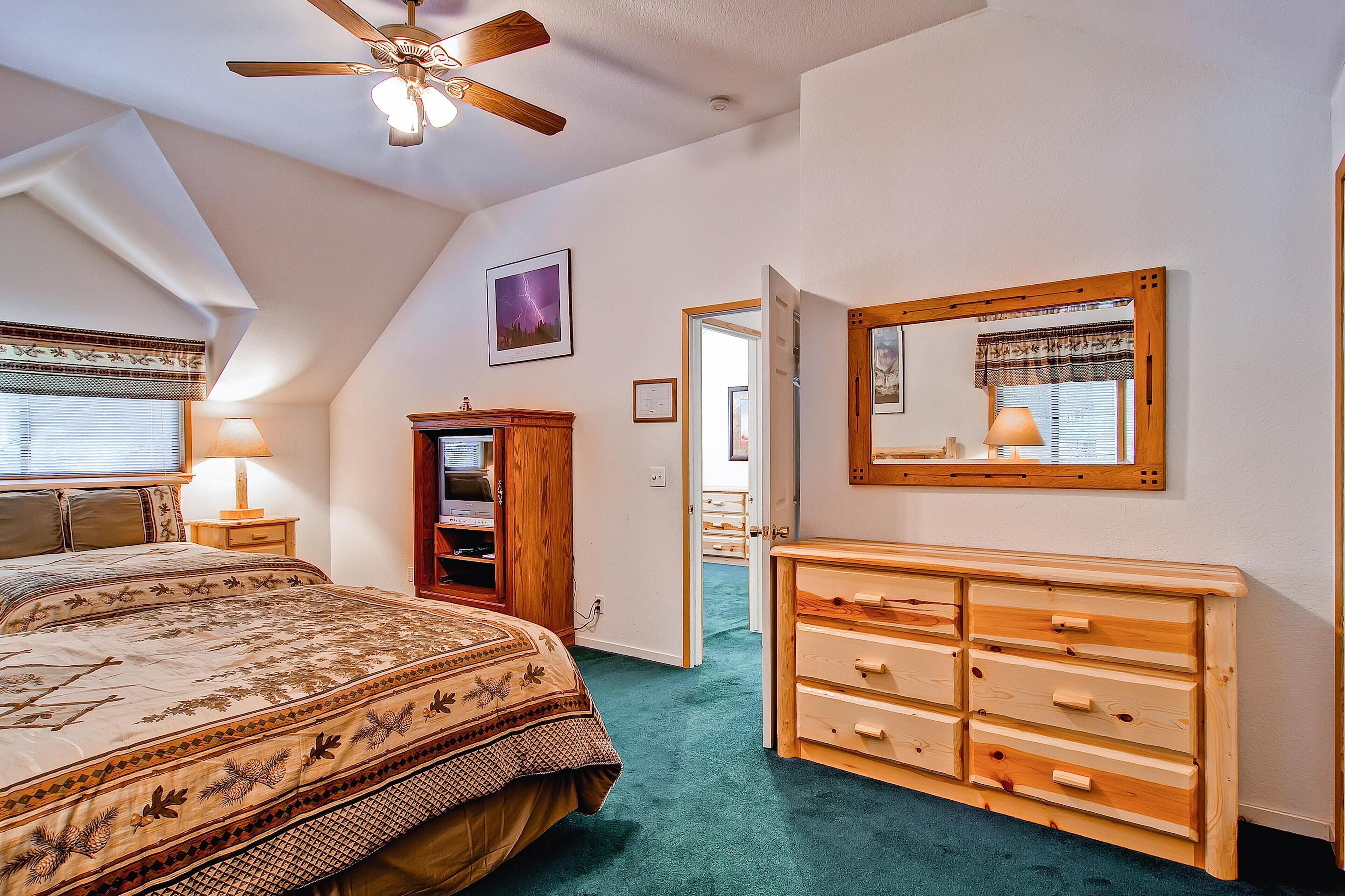 Une chambre à coucher comprenant un lit, une commode, un ventilateur de plafond, un miroir et une porte donnant sur une autre pièce.