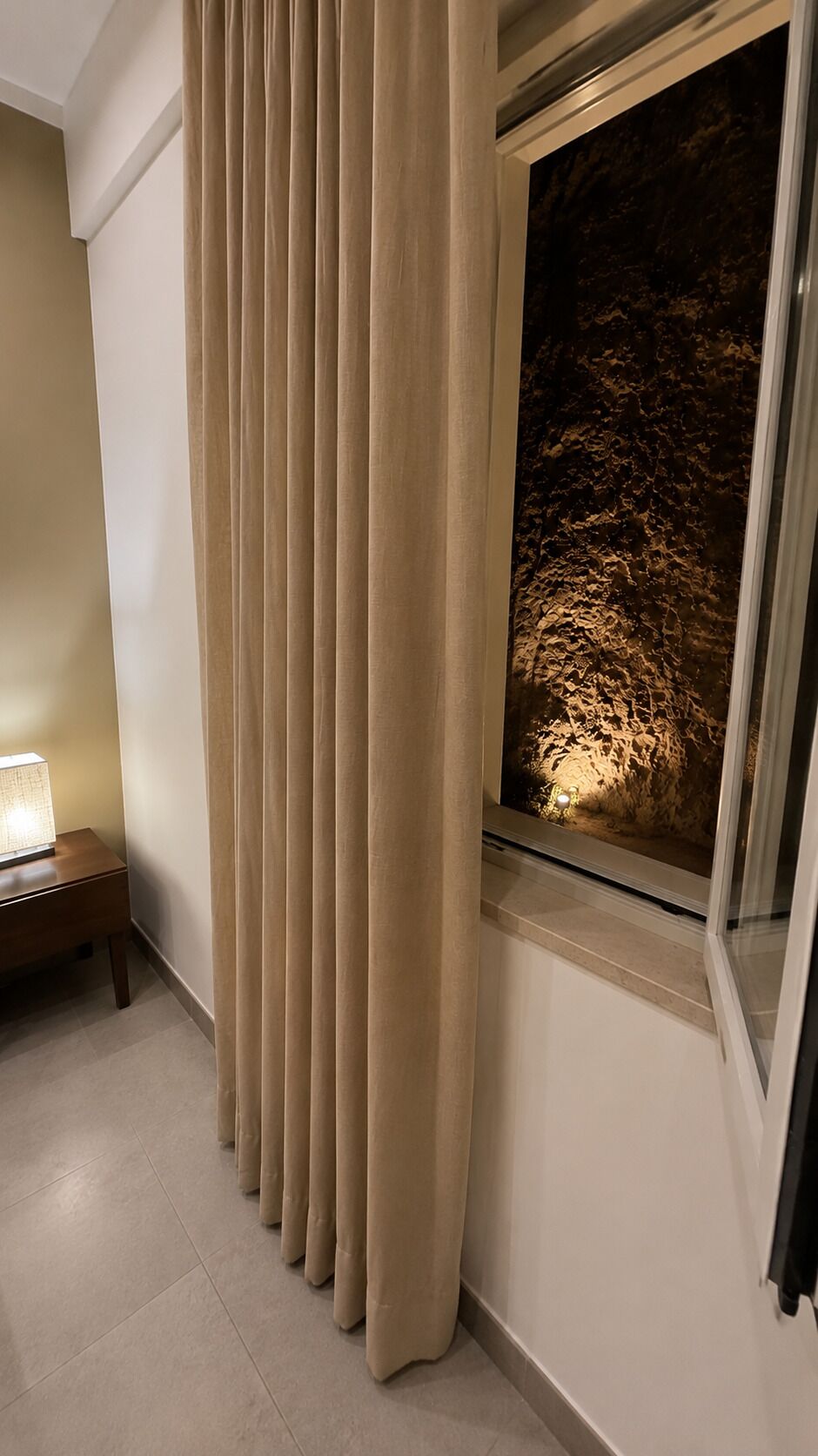 A room with a window, a lamp on a nightstand, and a curtain.