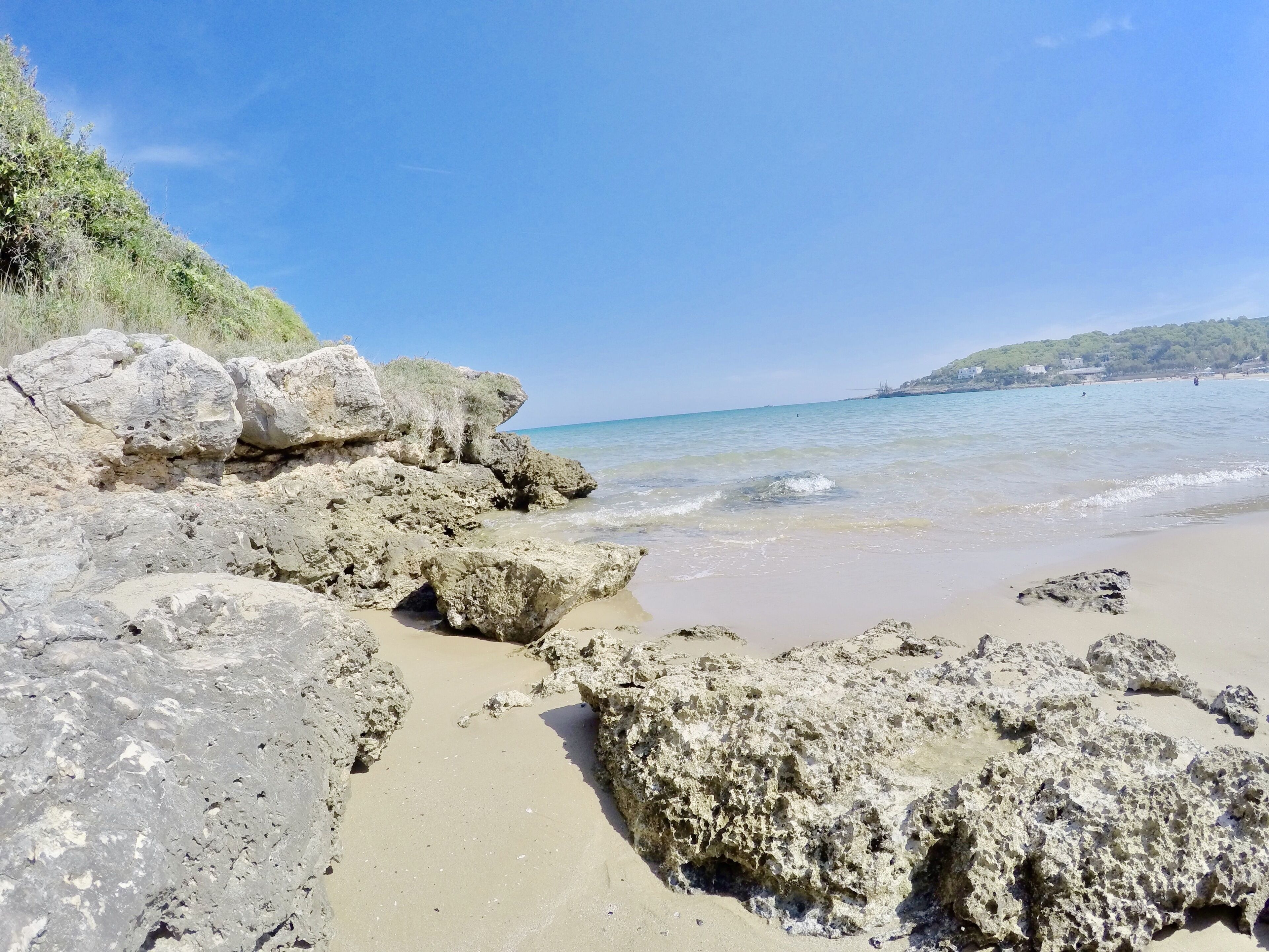 A rocky beach with clear blue water and a distant hill.
