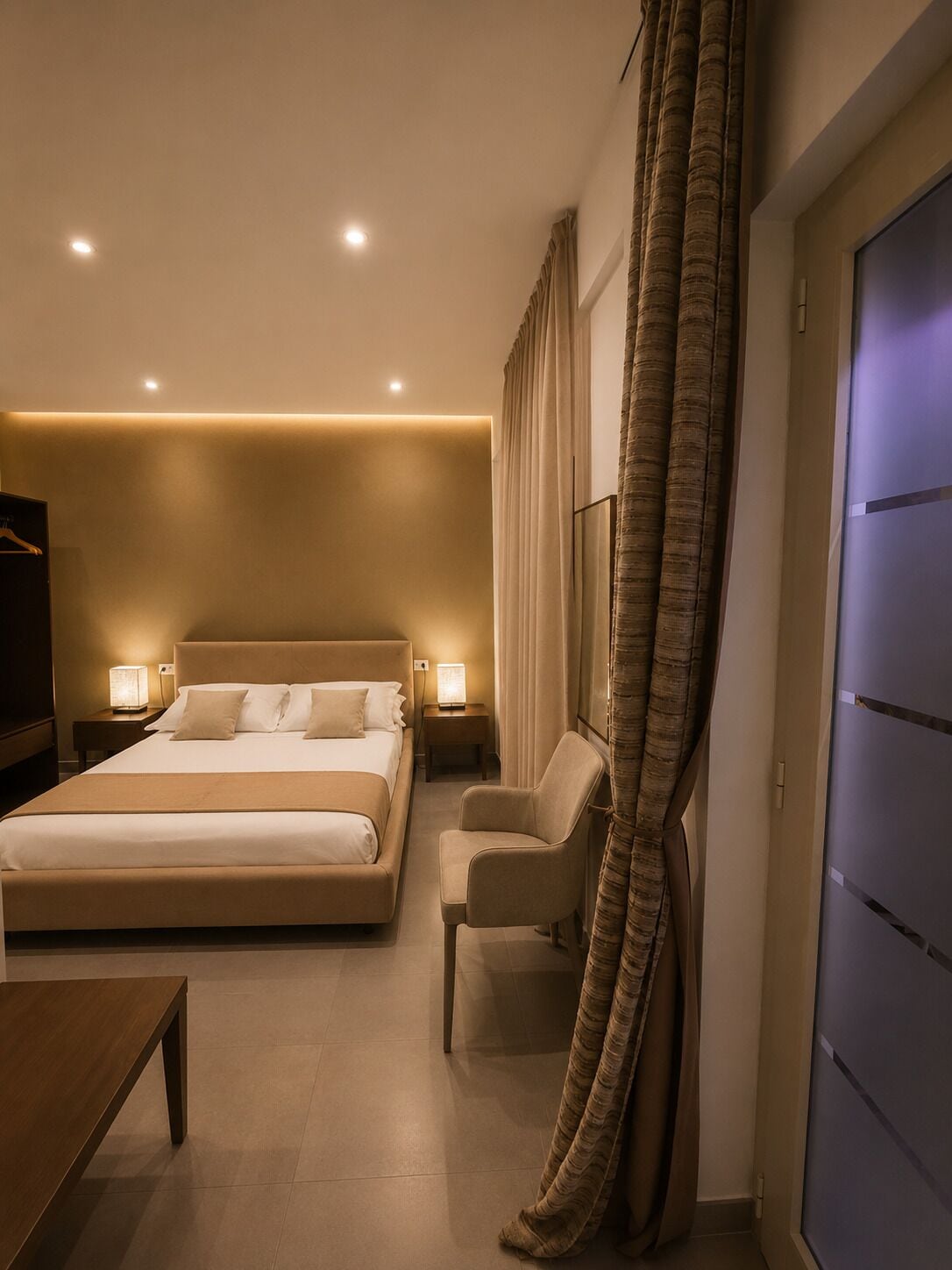 A hotel room with a large bed, bedside tables with lamps, a chair, a desk, and a wardrobe.