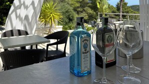 A table with two bottles of Bombay Sapphire gin and two empty wine glasses, set outdoors with a white pergola and greenery in the background. - Petra Maris B&B (Peschici)