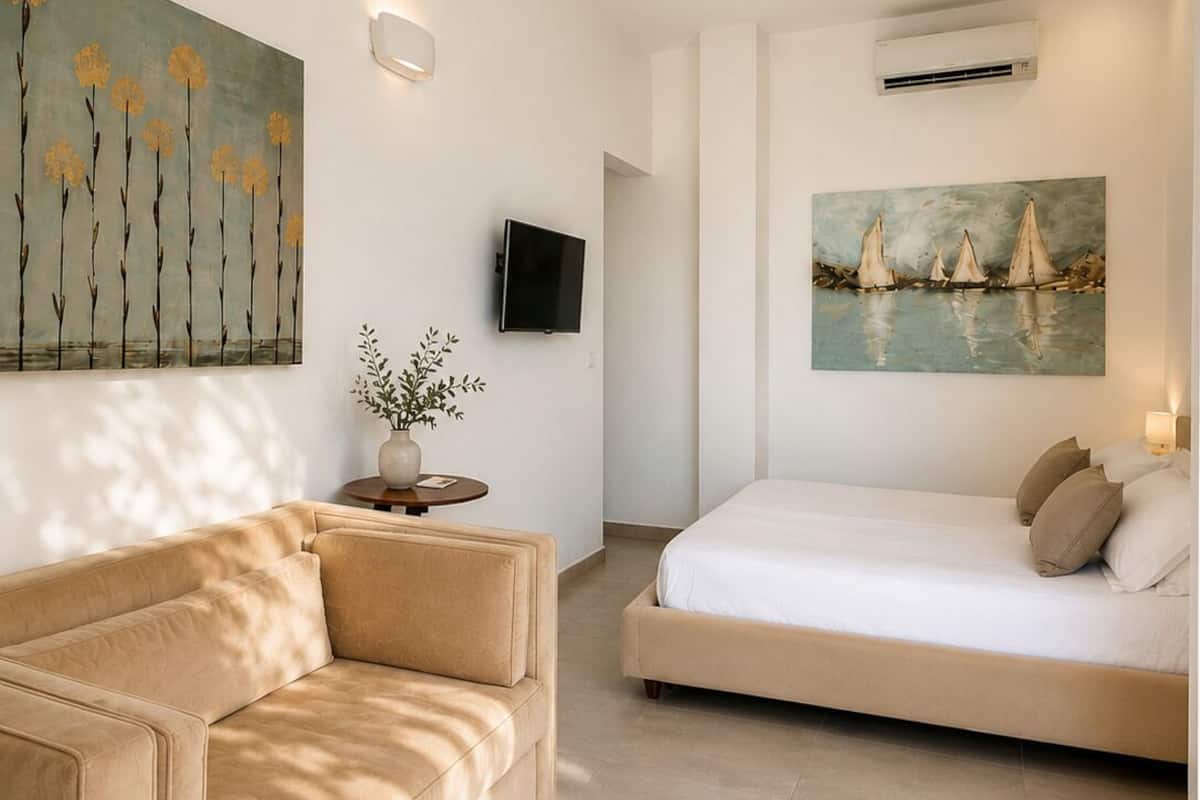 A modern hotel room with a sofa, a bed, a painting of sailboats, and a TV.