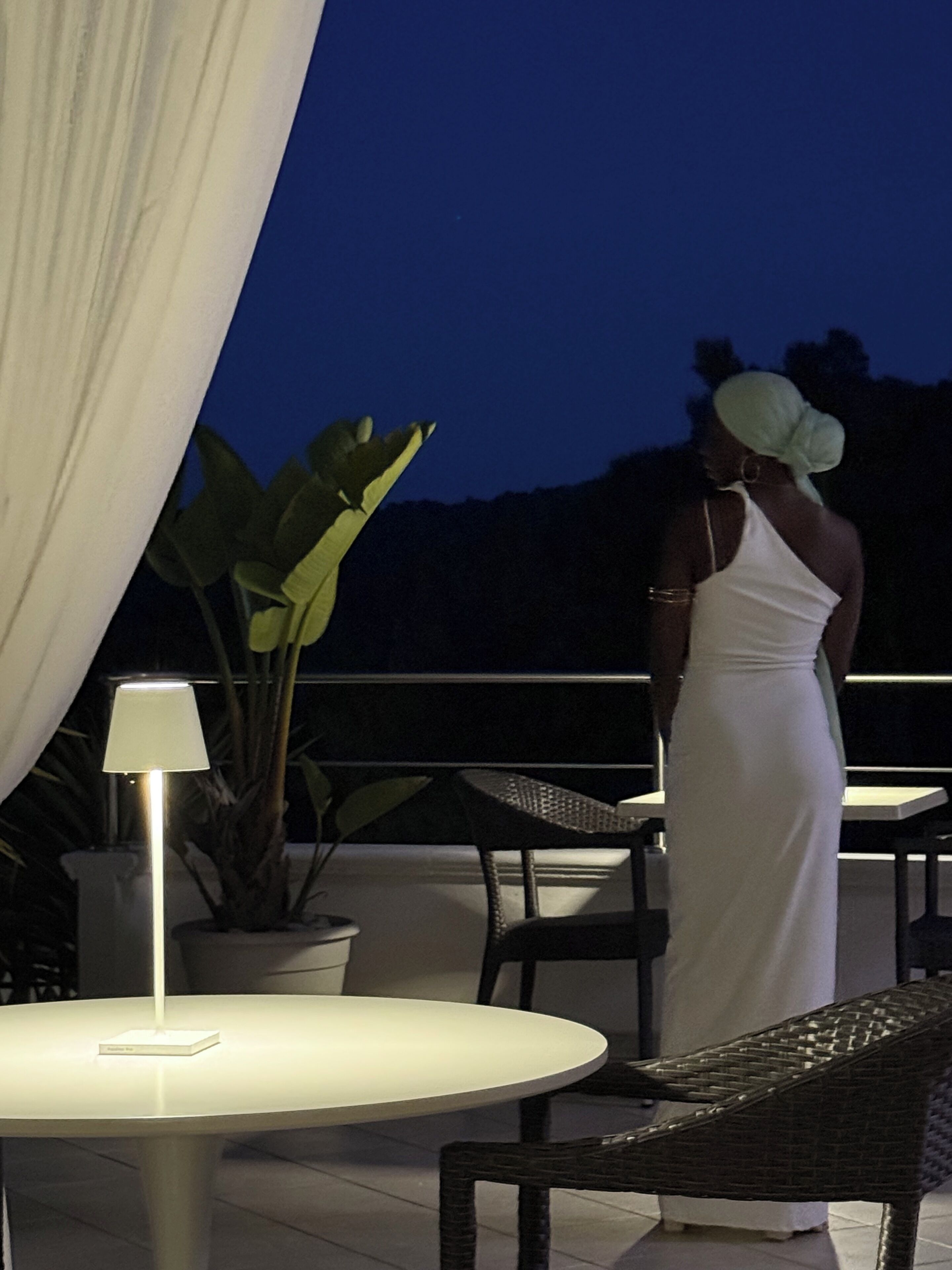 Una mujer con vestido blanco de pie junto a una mesa y sillas en un patio al aire libre de noche.