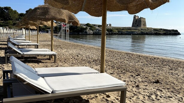 A beach with rows of sun loungers under thatched umbrellas, a rocky outcrop, and calm water in the background. - Petra Maris B&B (Peschici)