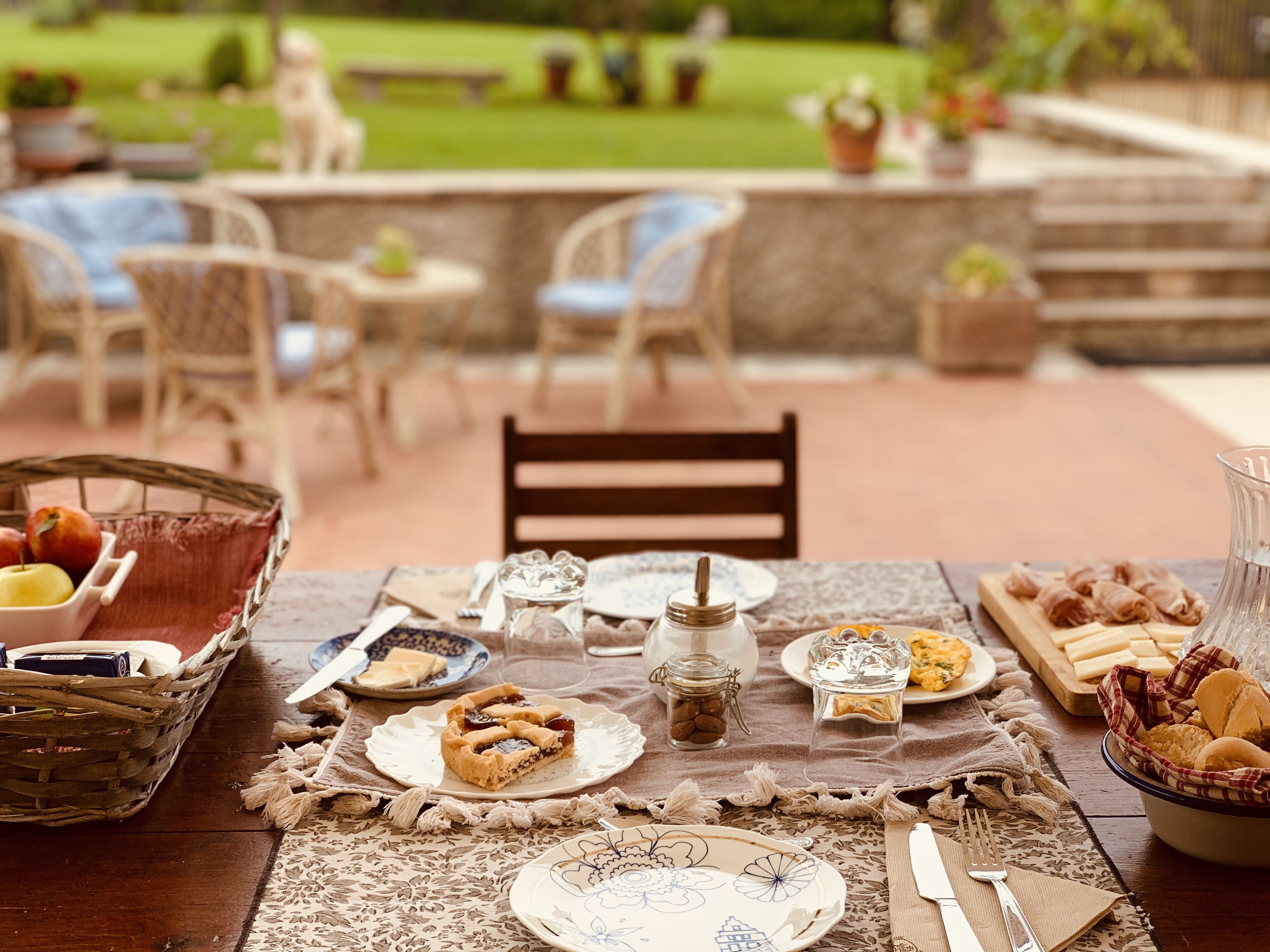 A dining table set outdoors with various dishes, a basket of fruits, and a view of a garden in the background.