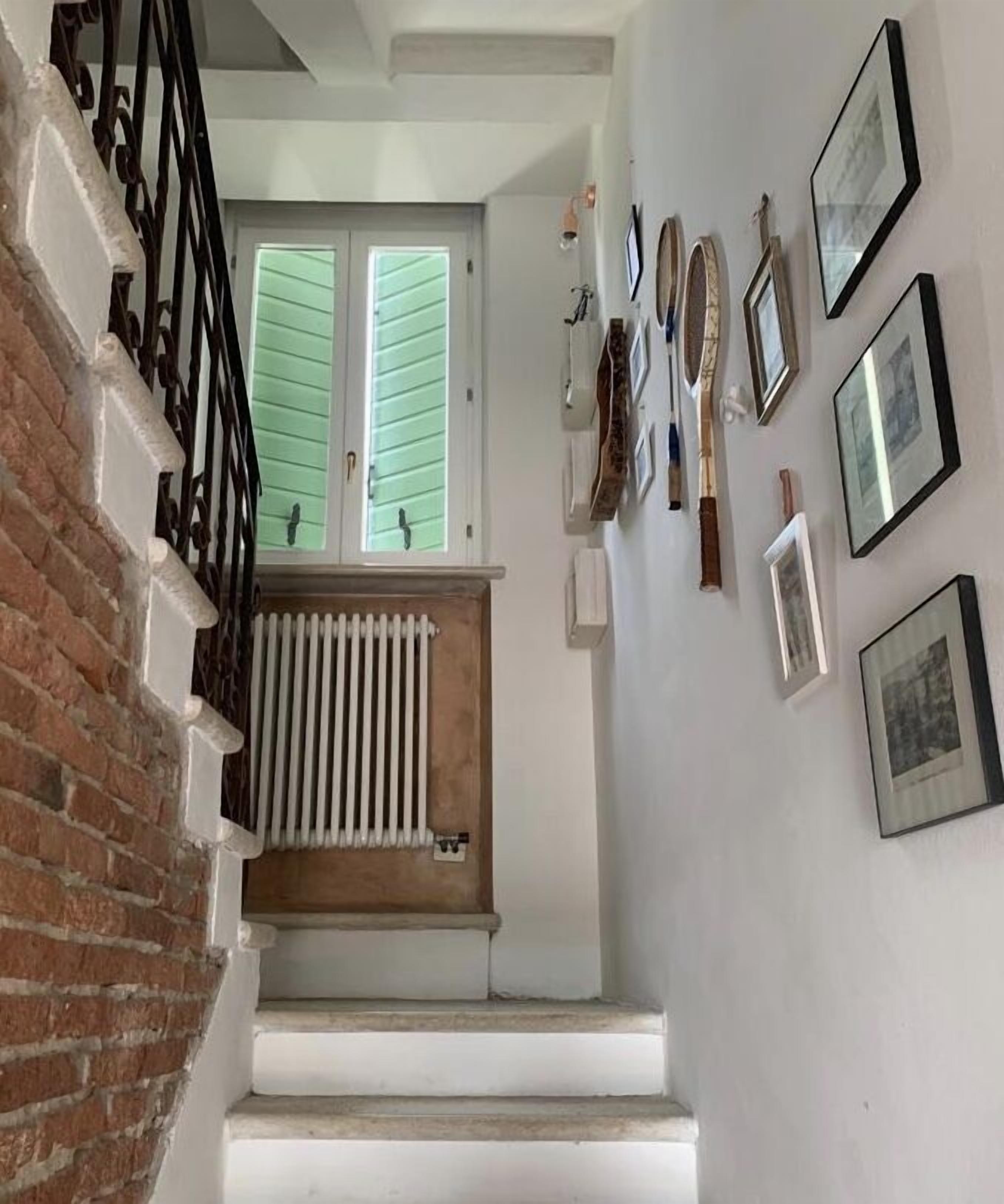 Un escalier, avec un mur de briques d’un côté et un mur blanc de l’autre, mène à une pièce équipée d’un radiateur et d’une fenêtre avec des volets.