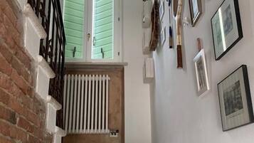 Un escalier, avec un mur de briques d’un côté et un mur blanc de l’autre, mène à une pièce équipée d’un radiateur et d’une fenêtre avec des volets.