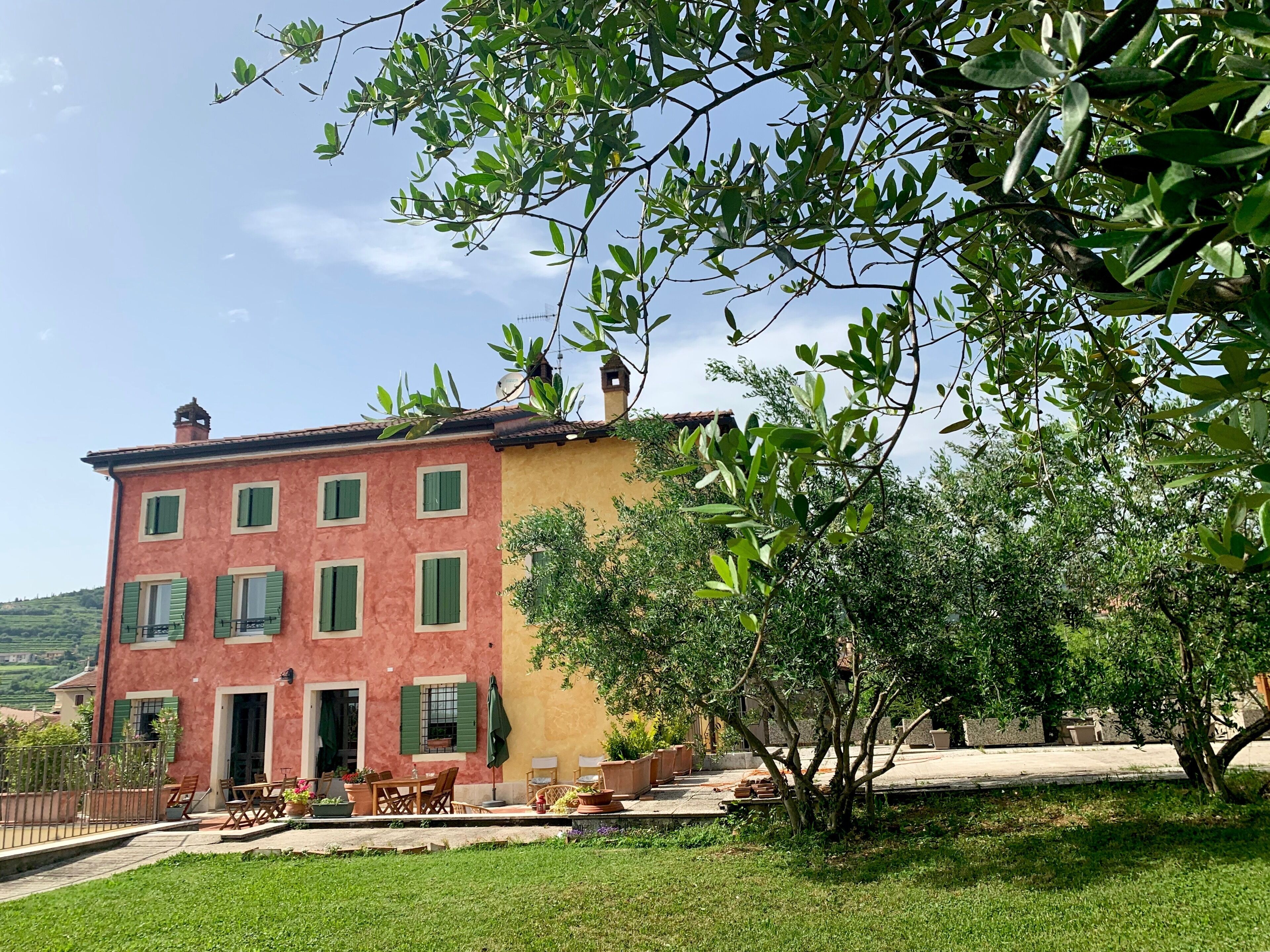 Un bâtiment à deux étages, doté d’une façade rouge et de volets verts, d’un petit porche avec des meubles d’extérieur, et offrant une vue sur la verdure environnante.