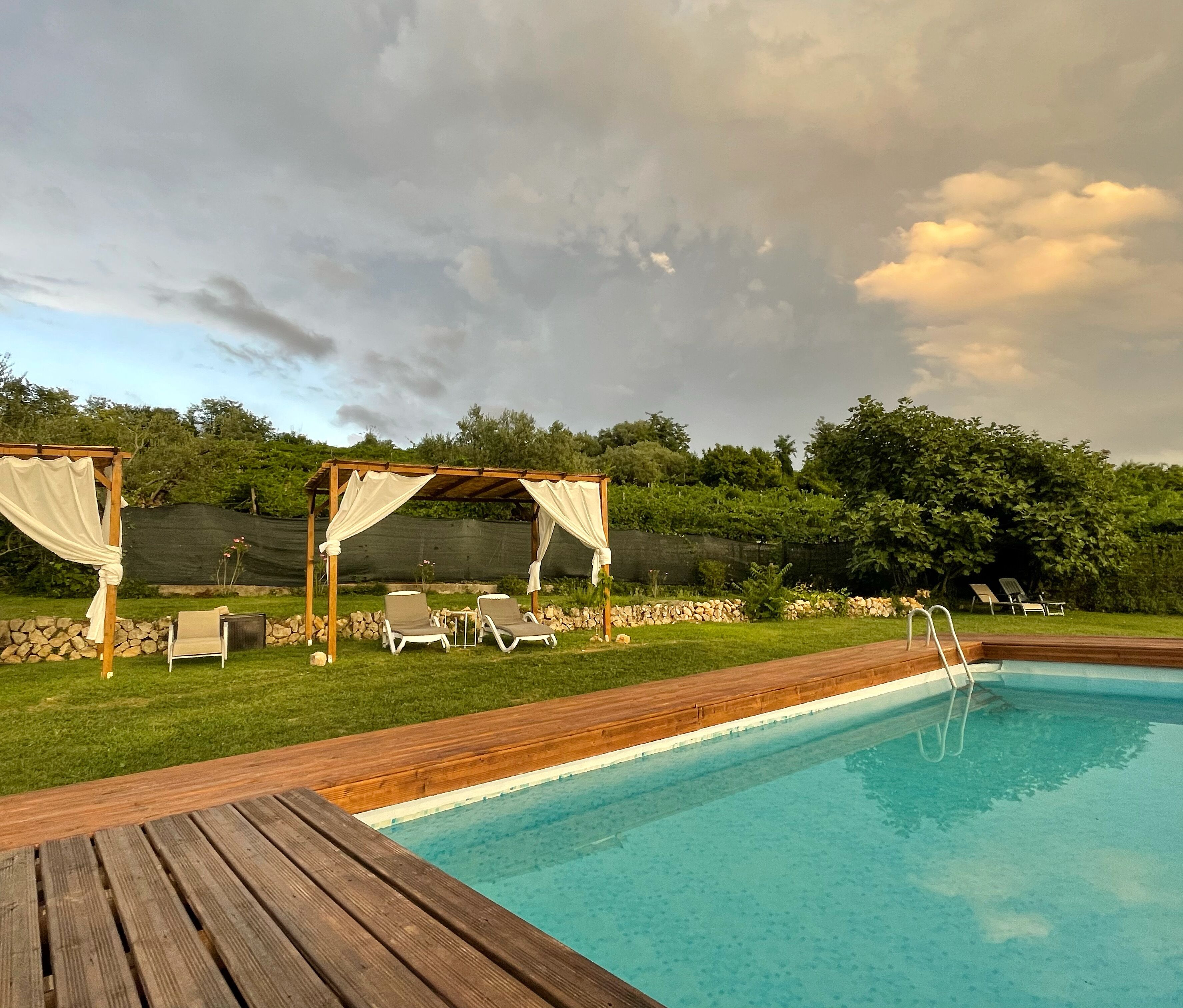Ein Schwimmbecken mit Holzterrasse, zwei Liegestühlen und einer Pergola mit Vorhängen.