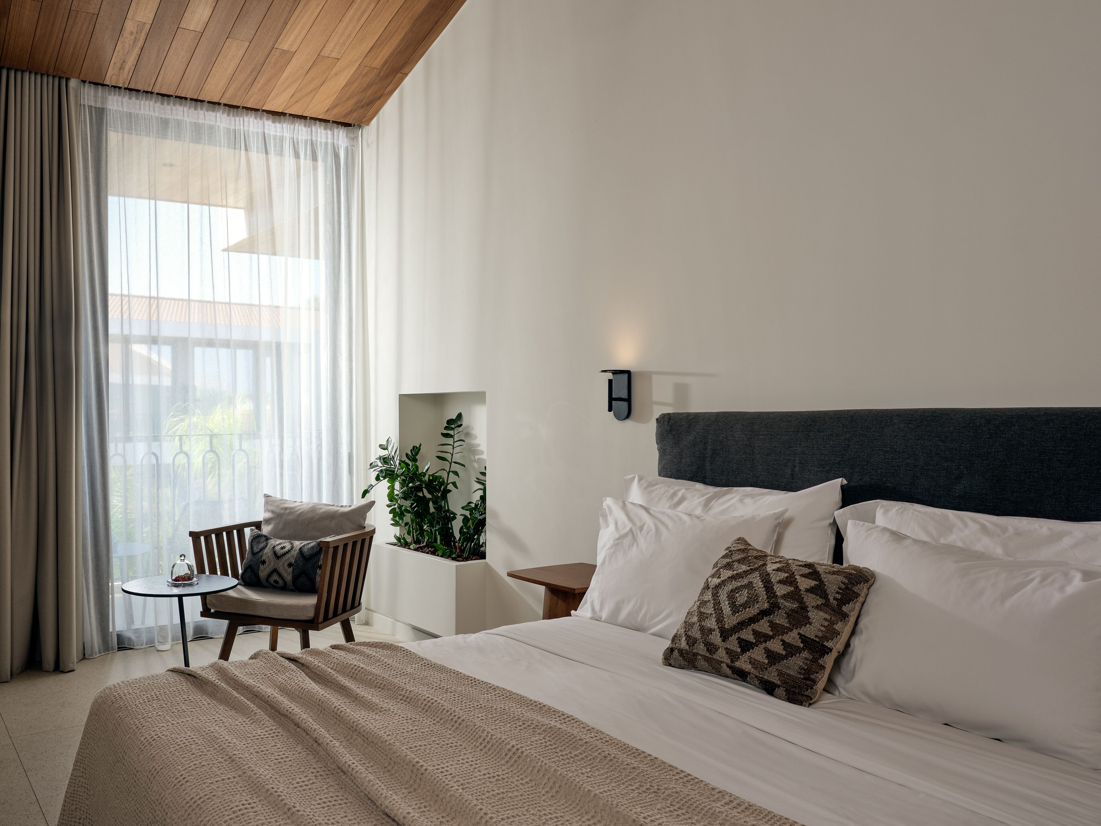 A modern hotel room with a large bed, a small table with a chair, a potted plant, and a view of greenery outside.