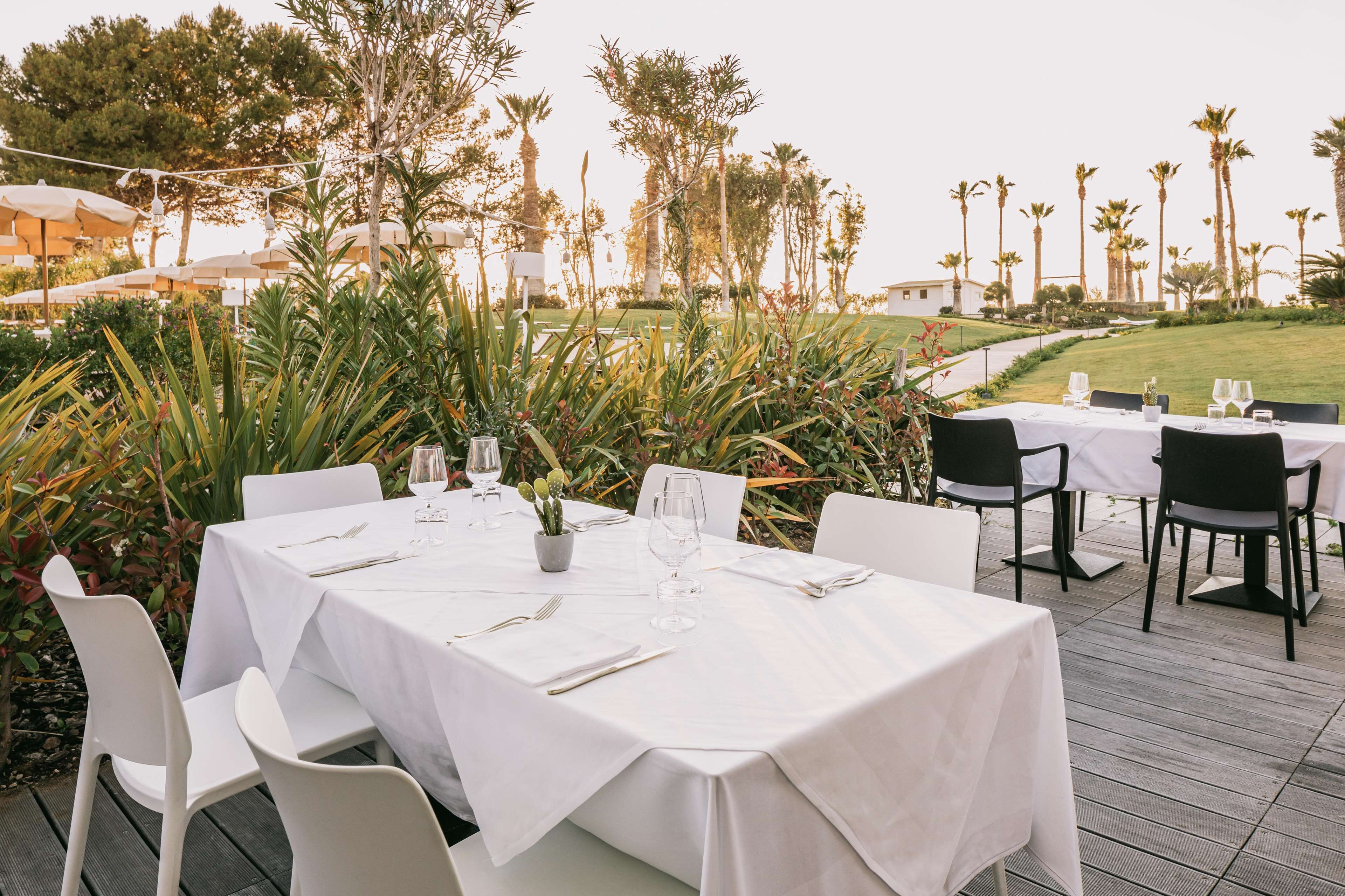 An outdoor dining area with white tablecloth-covered tables and chairs, surrounded by greenery and palm trees.