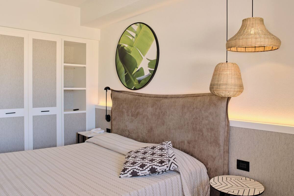 A bedroom with a bed, a headboard, a wall-mounted picture, a bedside table, and a lamp.