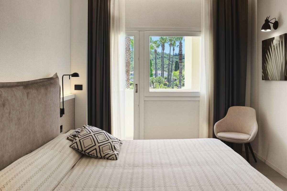 A hotel room with a bed, a chair, a window with a view of palm trees, and a wall-mounted lamp.