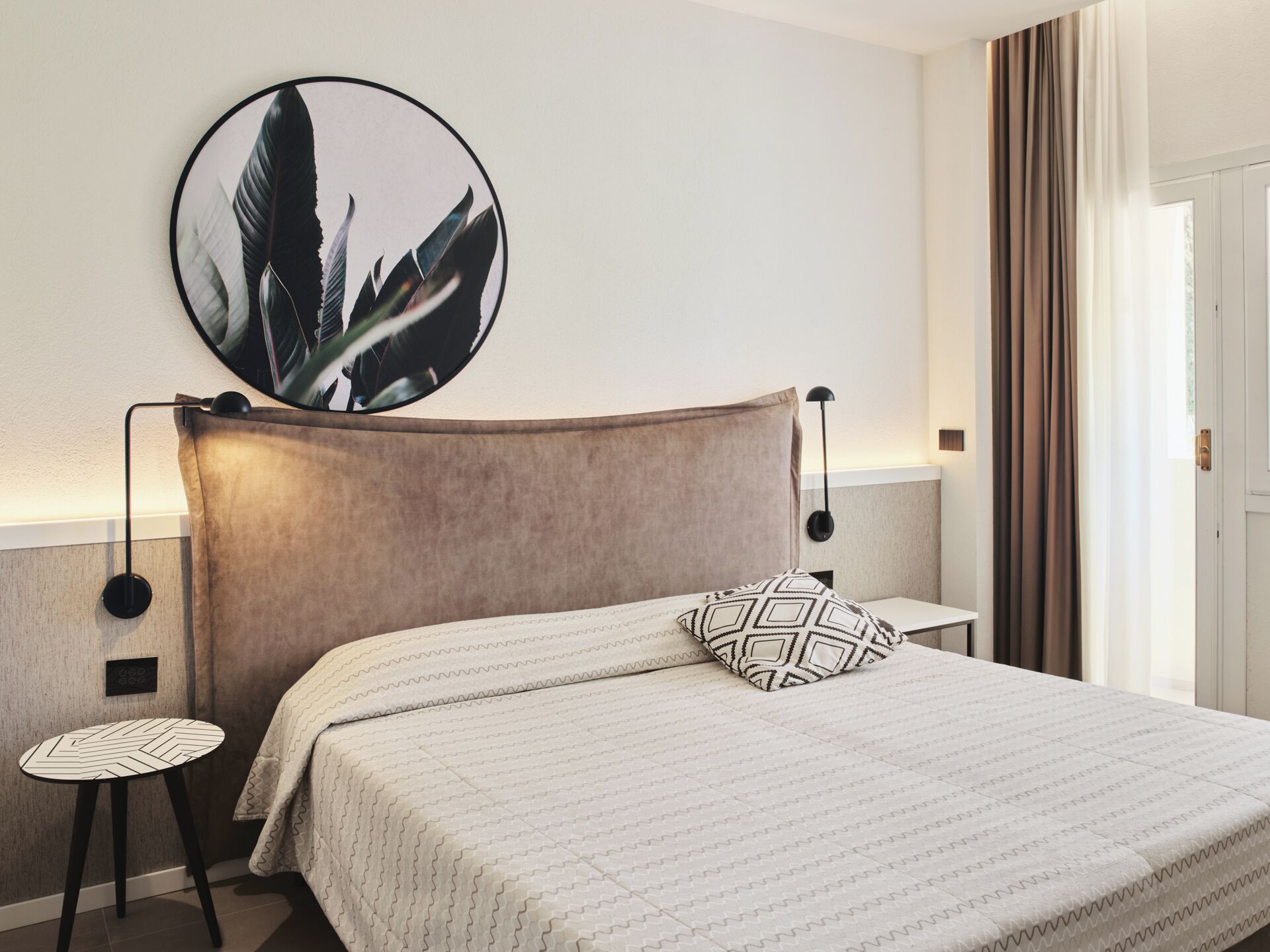 Habitación de hotel ordenada con una cama grande, una obra de arte redonda en la pared, una mesita auxiliar y vistas al exterior a través de una ventana.