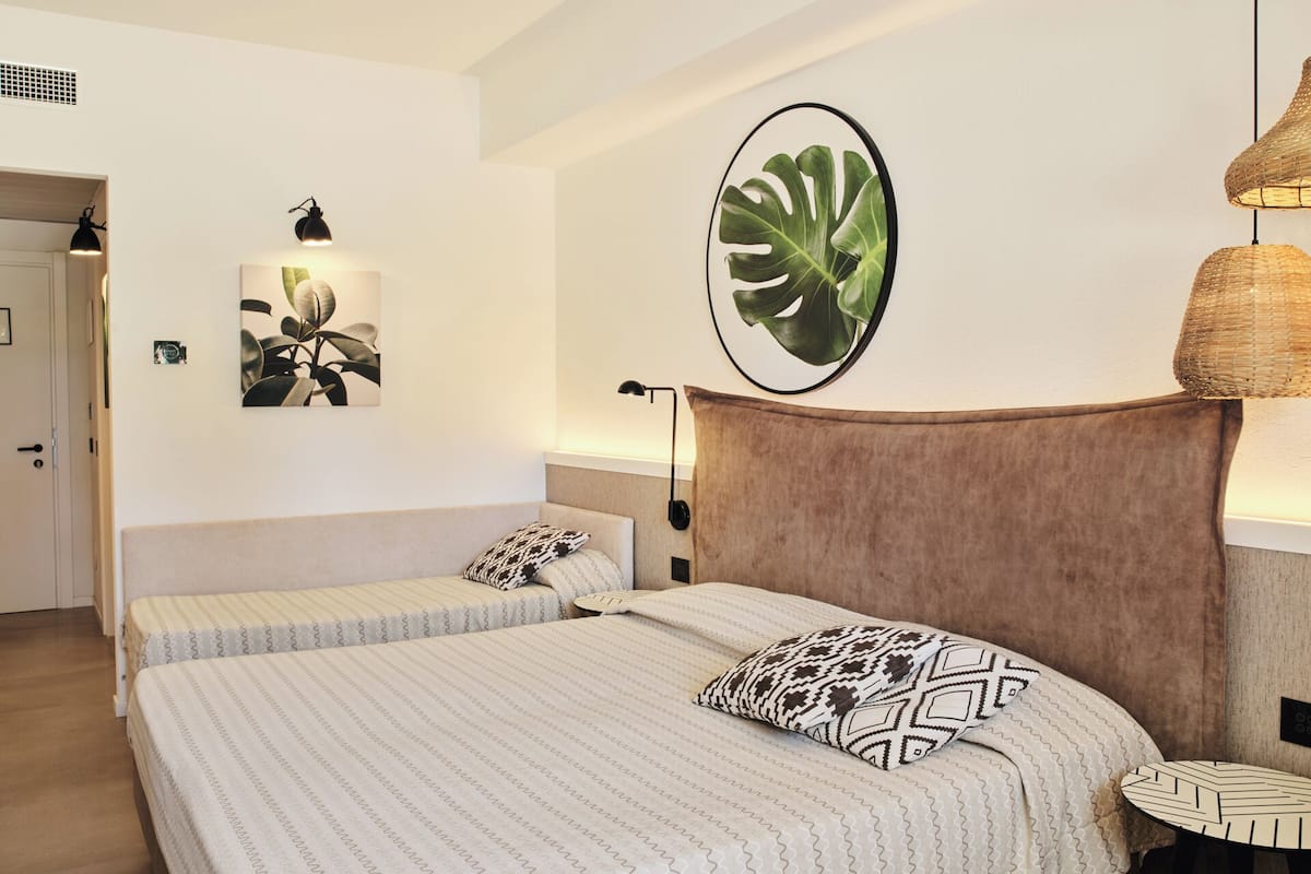 A hotel room with two beds, a large plant, a wall-mounted lamp, and a woven light fixture.