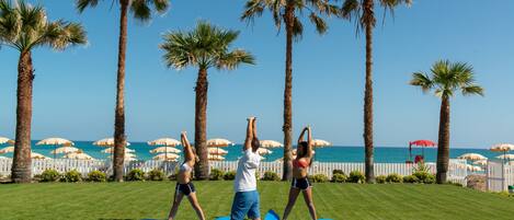 Tres personas practicando yoga sobre esterillas azules en un área cubierta de césped, con palmeras y sombrillas al fondo.