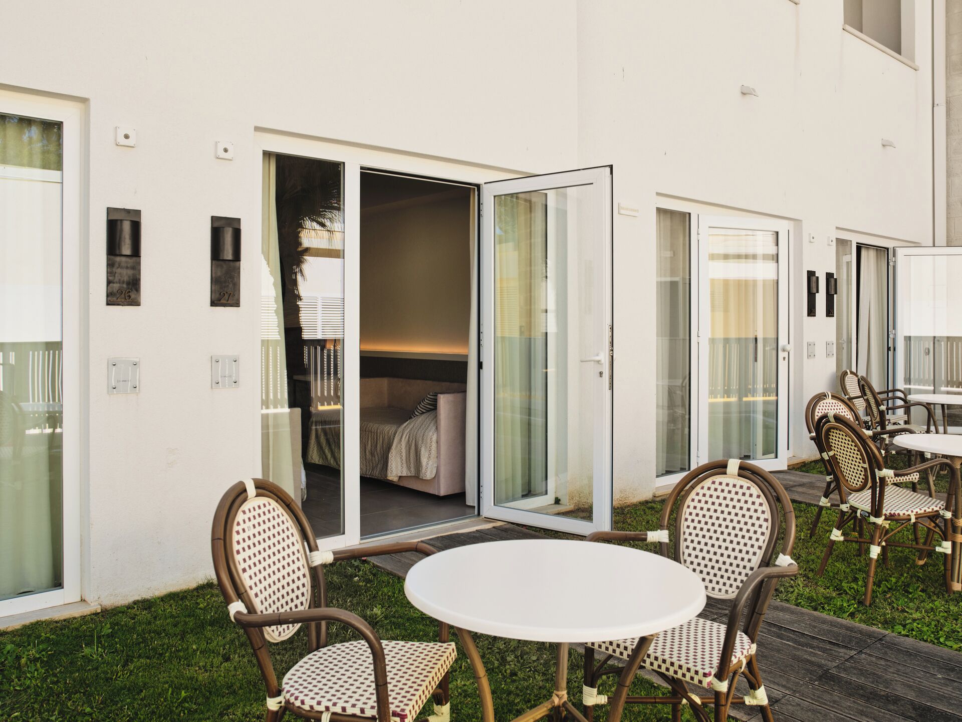 Une terrasse extérieure moderne, avec des chaises en osier et une table ronde, une porte-fenêtre coulissante donnant sur une chambre, et un mur extérieur blanc.