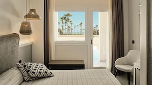 A hotel room with a bed, a bench, a chair, a window with a view of palm trees and a beach, and two hanging lamps. - Mangia's Himera Resort (Campofelice di Roccella)