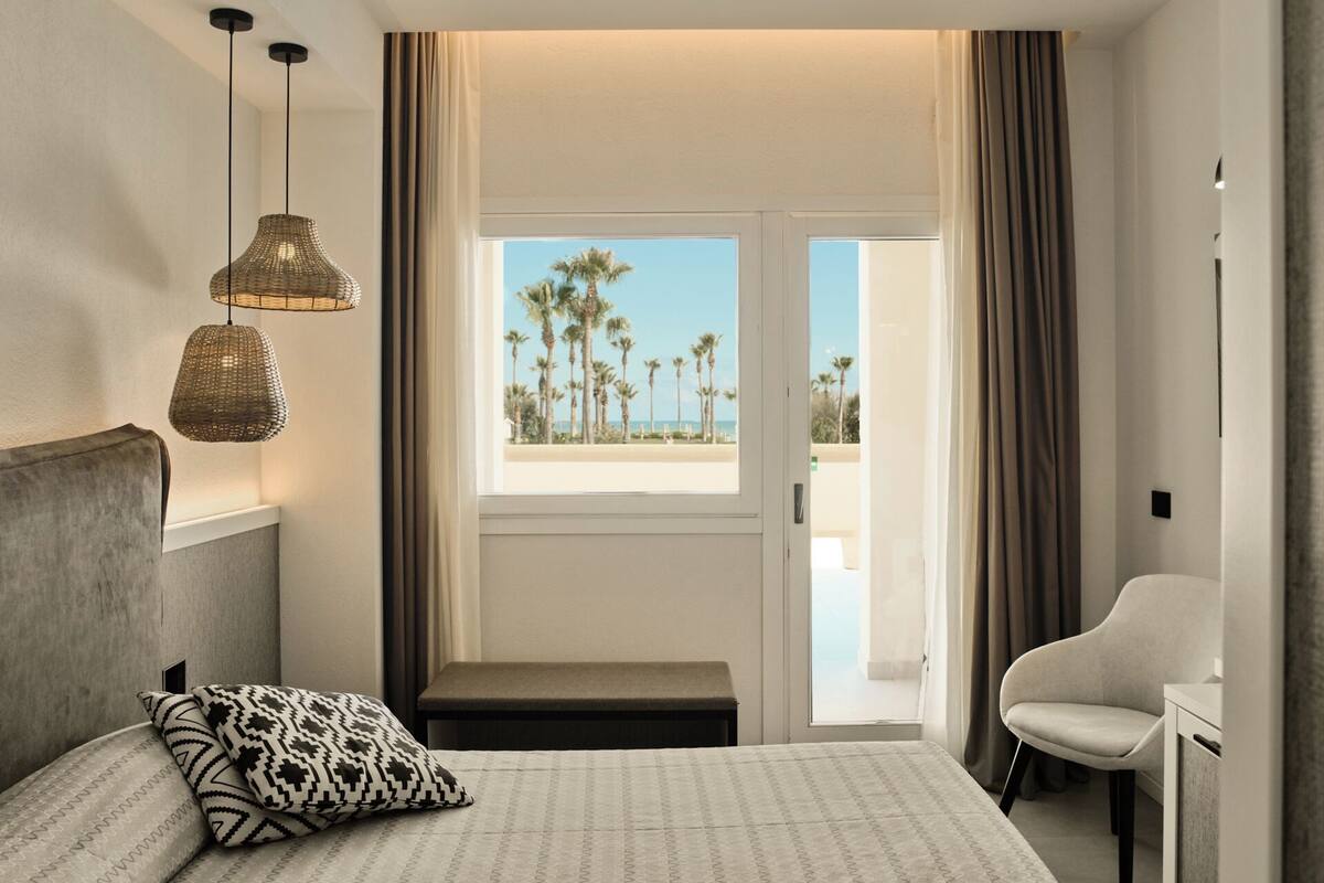 A hotel room with a bed, a bench, a chair, a window with a view of palm trees and a beach, and two hanging lamps.