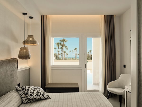 Une chambre d’hôtel comprenant un lit, une banquette, une chaise, une fenêtre donnant sur des palmiers et une plage, ainsi que deux lampes suspendues.