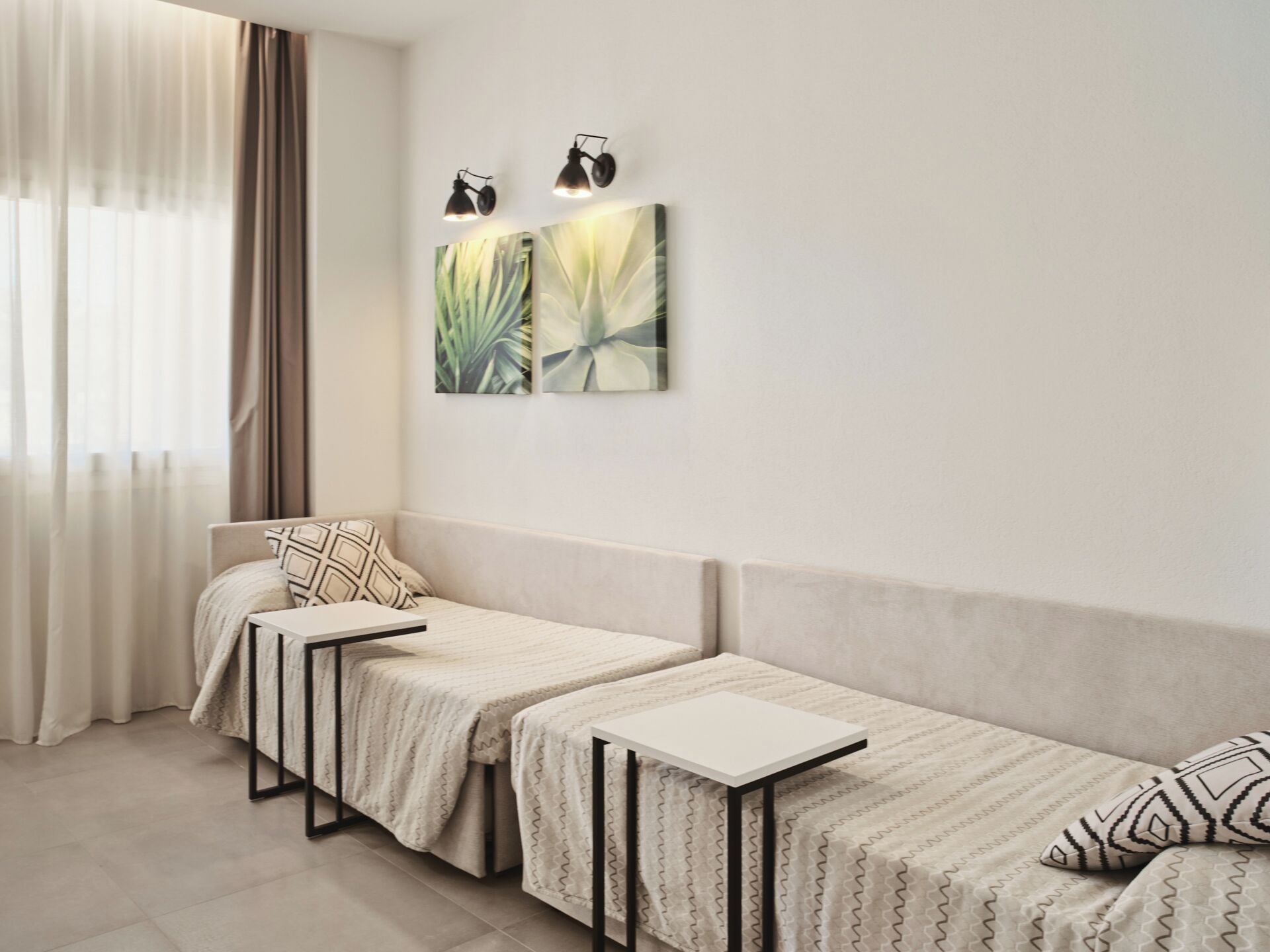 A room with a bed, a small table, and a bench. There are two wall-mounted lights and two paintings.