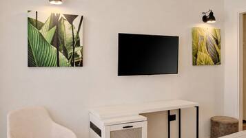 A room with a bed, a white desk, a chair, a flat-screen TV, and two wall-mounted lamps.