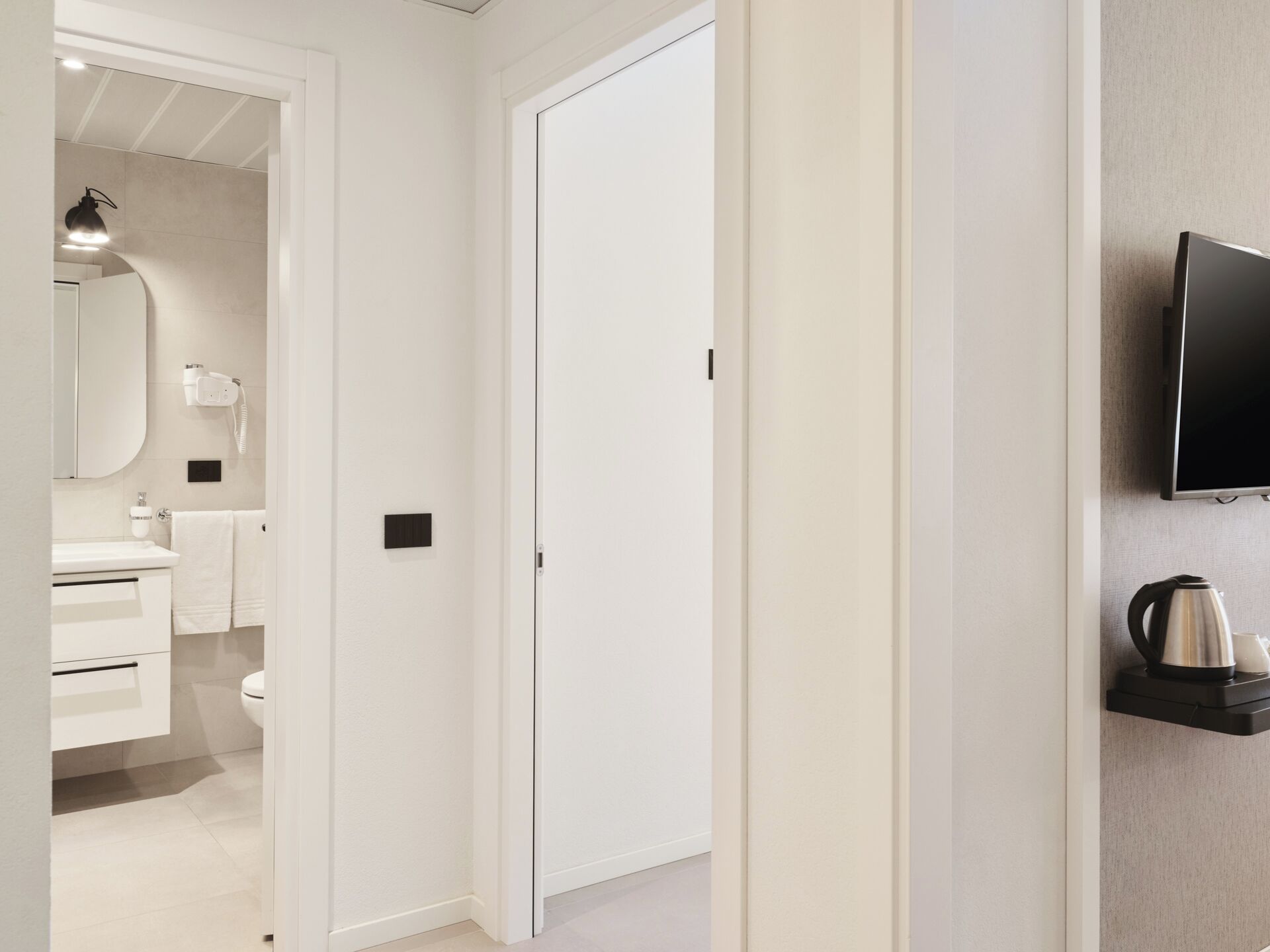 A hallway leading to a bathroom with a toilet, sink, and towel rack. A television is mounted on the wall, and a kettle is on a shelf.