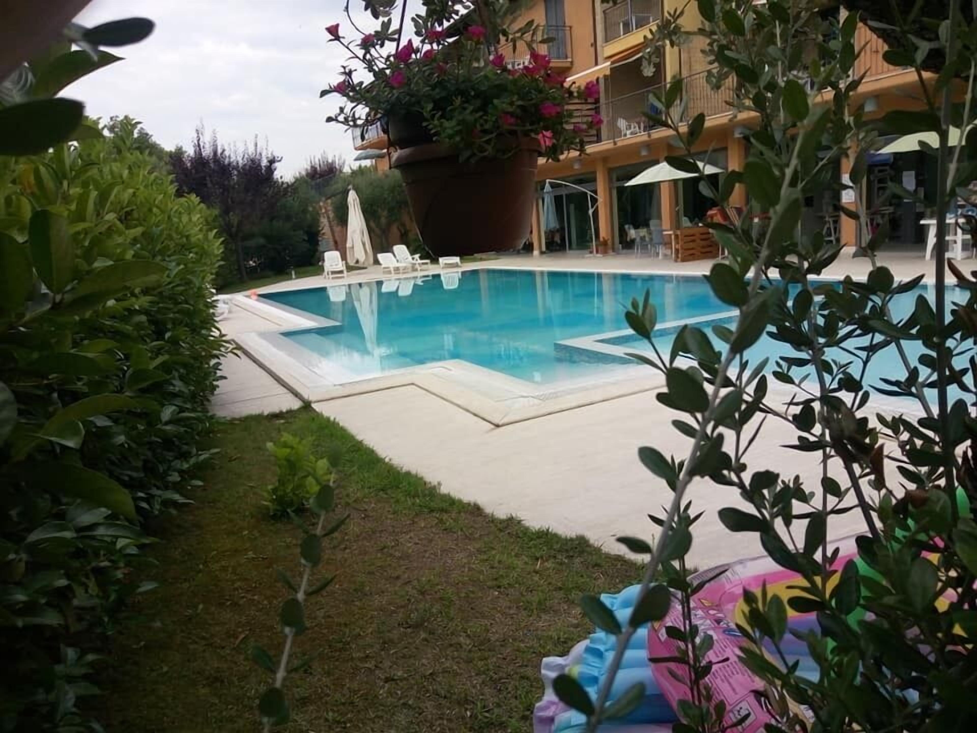 Una piscina amb gandules, envoltada de vegetació i un edifici amb balcons.