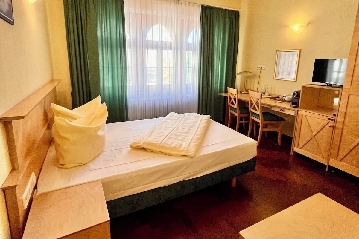 Ein Hotelzimmer mit Bett, Schreibtisch, Stuhl, Fernseher und einem Fenster mit Vorhängen.