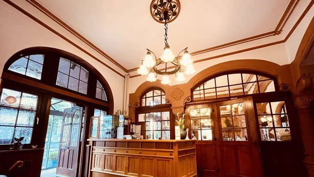 A spacious lobby with a central wooden reception desk, large arched windows, and a decorative ceiling light fixture. - Hotel Hoepfner Burghof (Karlsruhe)