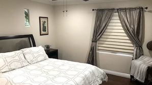 Ein Schlafzimmer mit Bett, Nachttisch, Sessel, Fenster mit Vorhängen, Deckenventilator und einem gerahmten Bild an der Wand.
