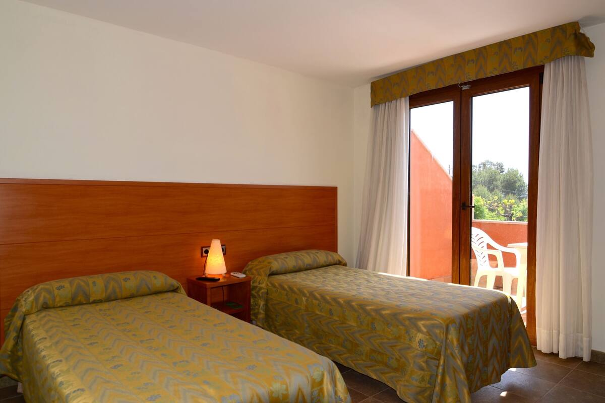 Habitación de hotel con dos camas, cabecera de madera, mesita de noche con lámpara y vista a un edificio rojo a través de una puerta de vidrio.