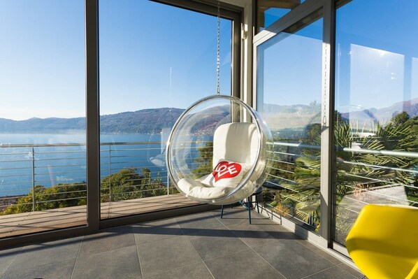 A clear view of a balcony with a hanging chair and a yellow chair, overlooking a body of water and mountains.