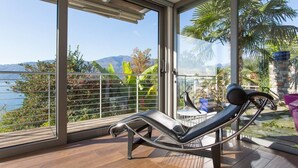 A balcony with a reclining chair, glass doors, and a view of the sea and mountains. - Luxury Italian Lakes Villa With Private Pool, Gym, Bbq, Wifi, Lake Views (Verbania)