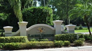 Un letrero con el nombre 'Laurel Greens' y un escudo, rodeado de setos cuidados y palmeras.