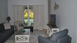 Una sala de estar con sofá, sillón, mesa de centro, televisor y vistas a través de una ventana con cortinas.