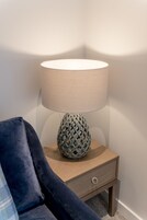 Ett litet bord som står i ett hörn vid sängen, med en lampa, en låda och en blå stol.