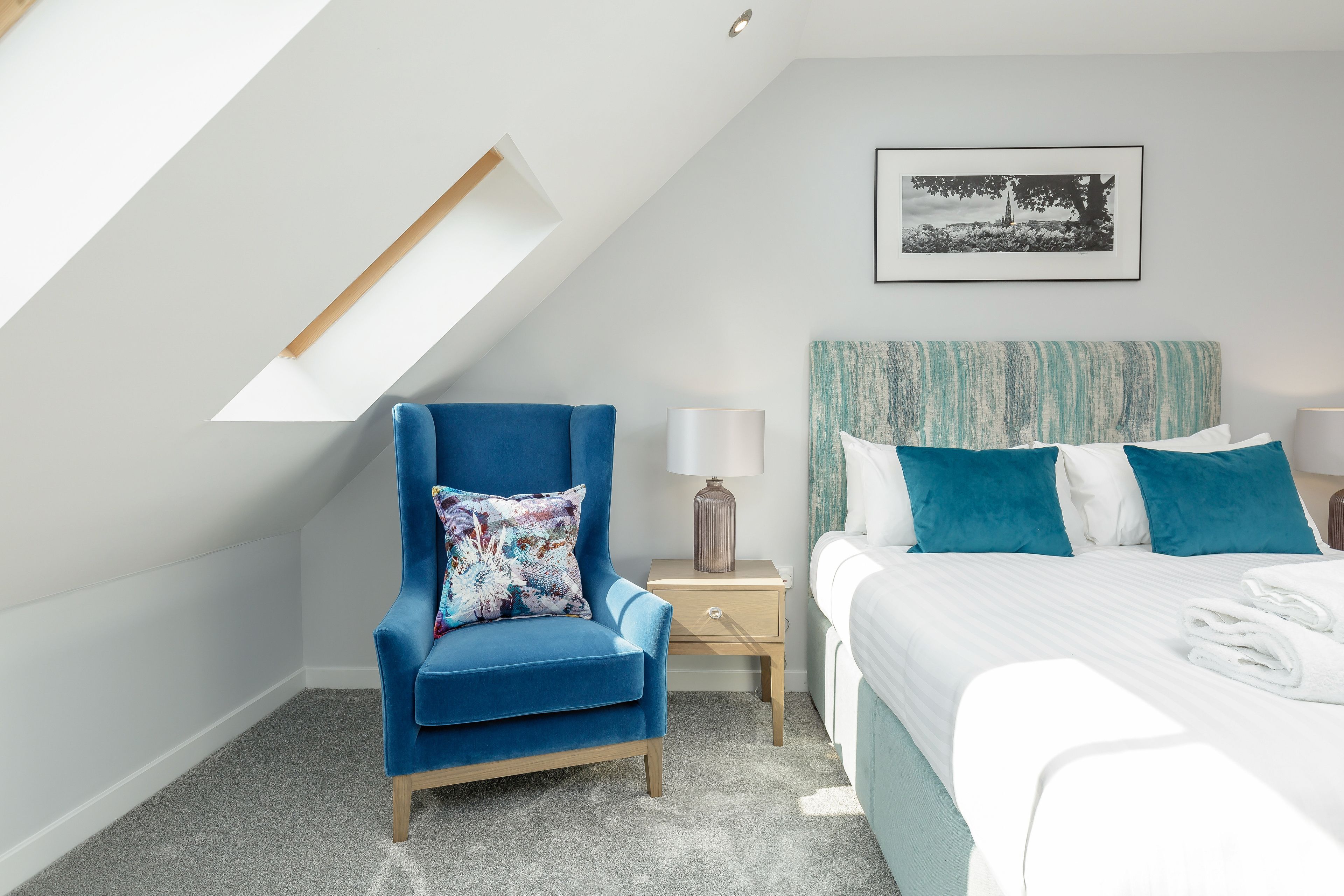 A bedroom with a sloped ceiling, a blue armchair, a bed with blue and white bedding, a nightstand with a lamp, and a framed picture on the wall.