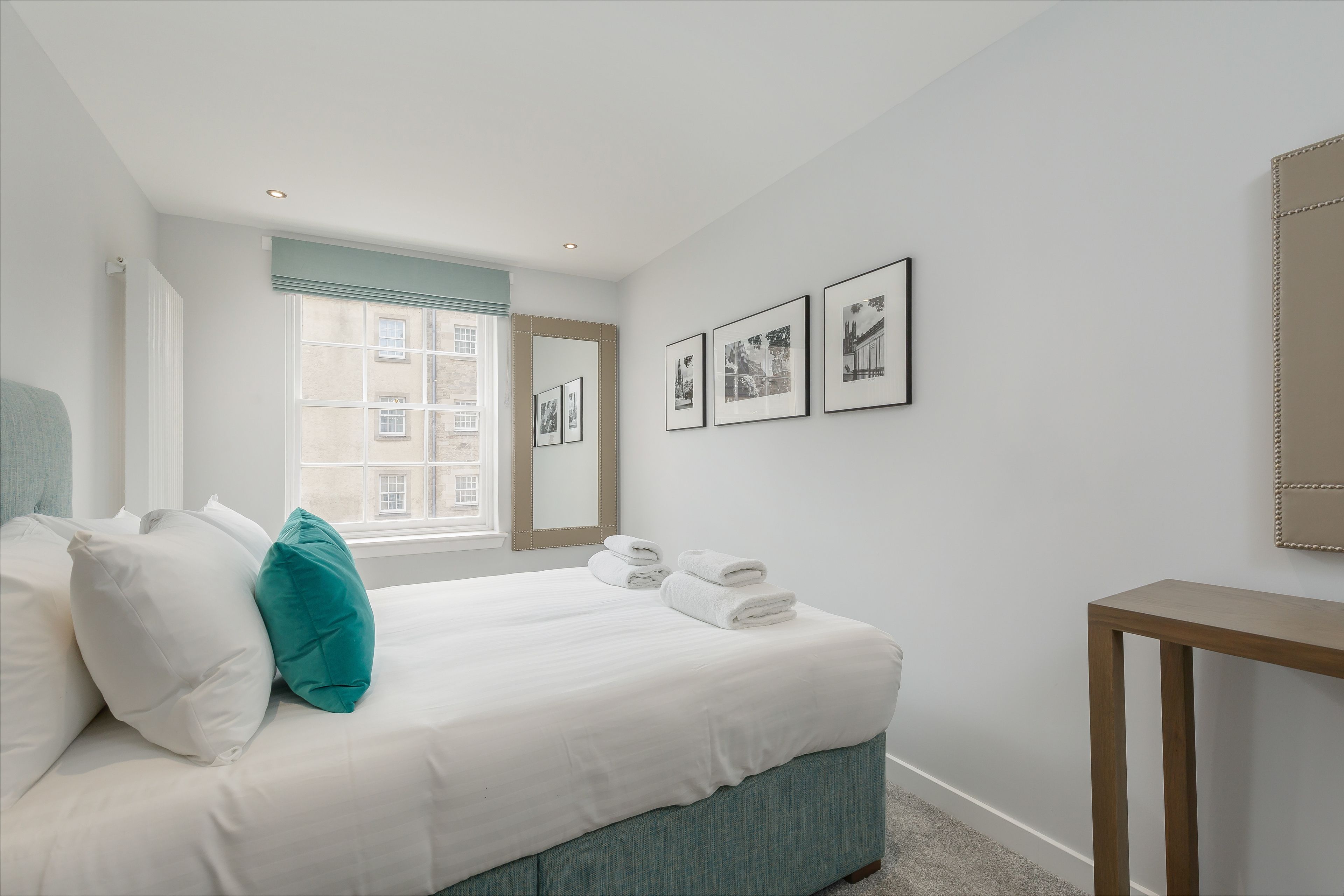 A neatly made bed with white linens and teal pillows, a bedside table, and framed pictures on the wall.