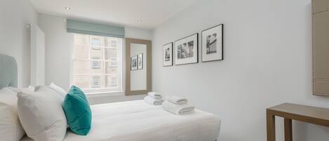 Ein ordentlich bezogenes Bett mit weißen Leinen und Türkis-Kissen, ein Nachttisch und gerahmte Bilder an der Wand.