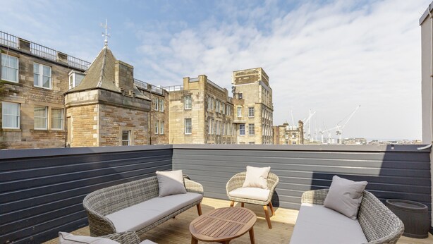 A rooftop terrace with wicker furniture, a round table, and a view of historic buildings and cranes in the background. - Destiny Scotland Royal Mile Residence (Edinburgh)