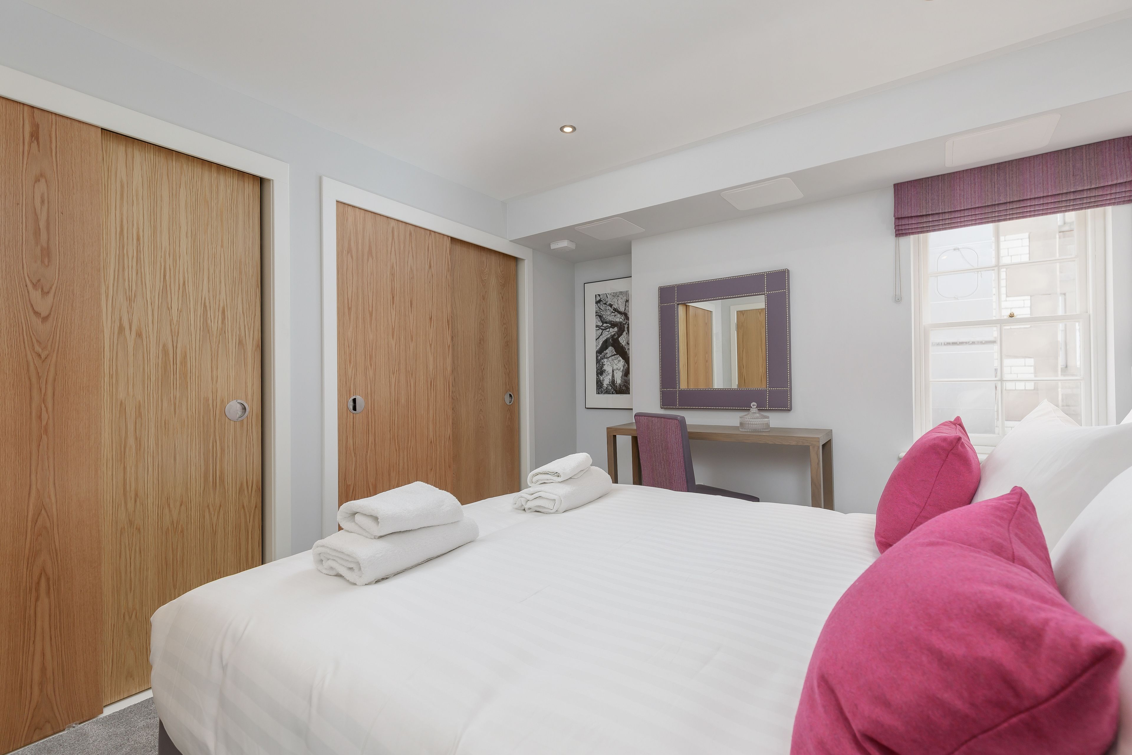 A hotel room with a large bed, a wooden wardrobe, a desk with a chair, a mirror, and a window with curtains.
