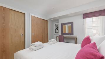 A hotel room with a large bed, a wooden wardrobe, a desk with a chair, a mirror, and a window with curtains.