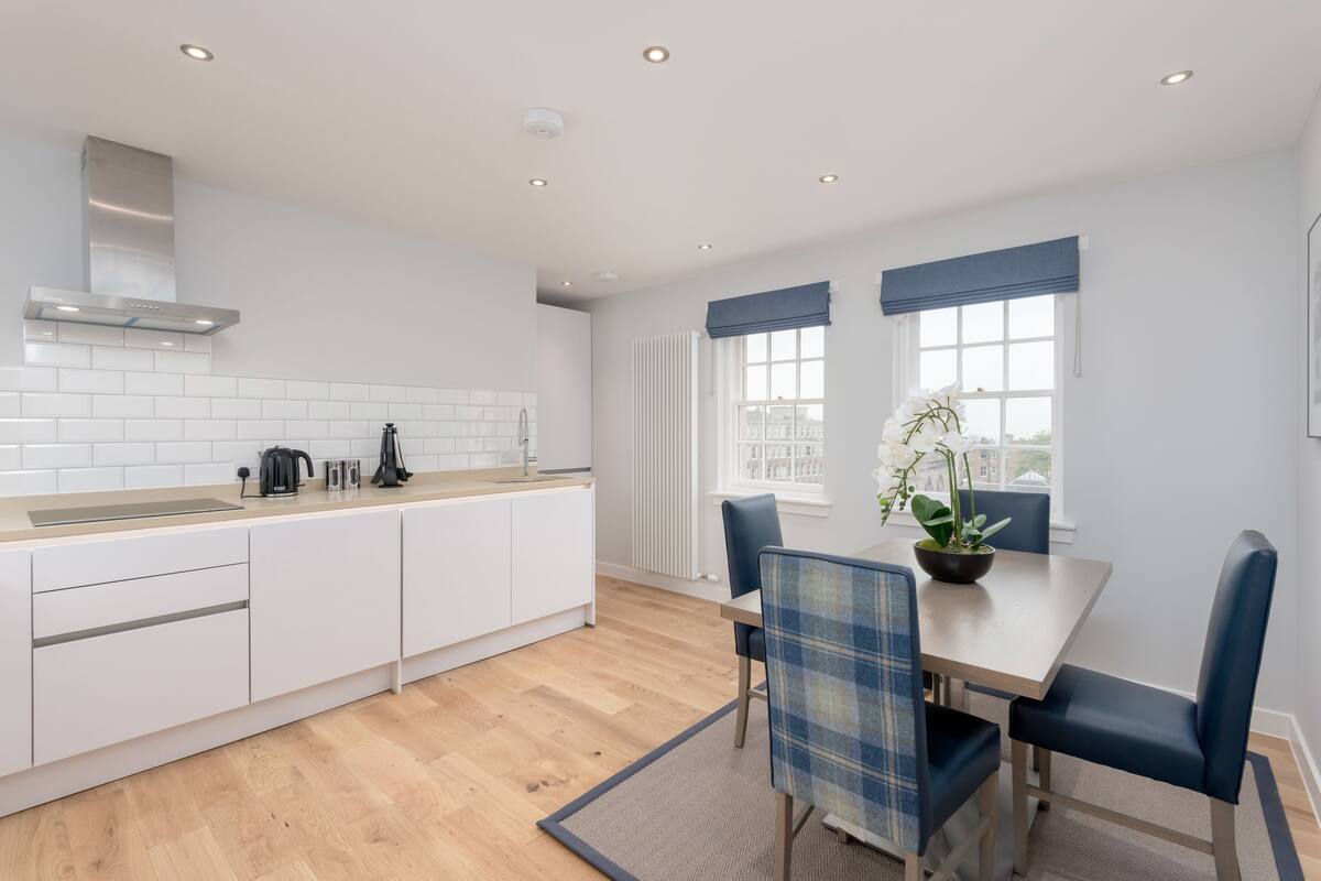 A modern kitchen with white cabinets, a wooden countertop, and a built-in stove. There is a dining area with a table and chairs, and a window with a view of the city.