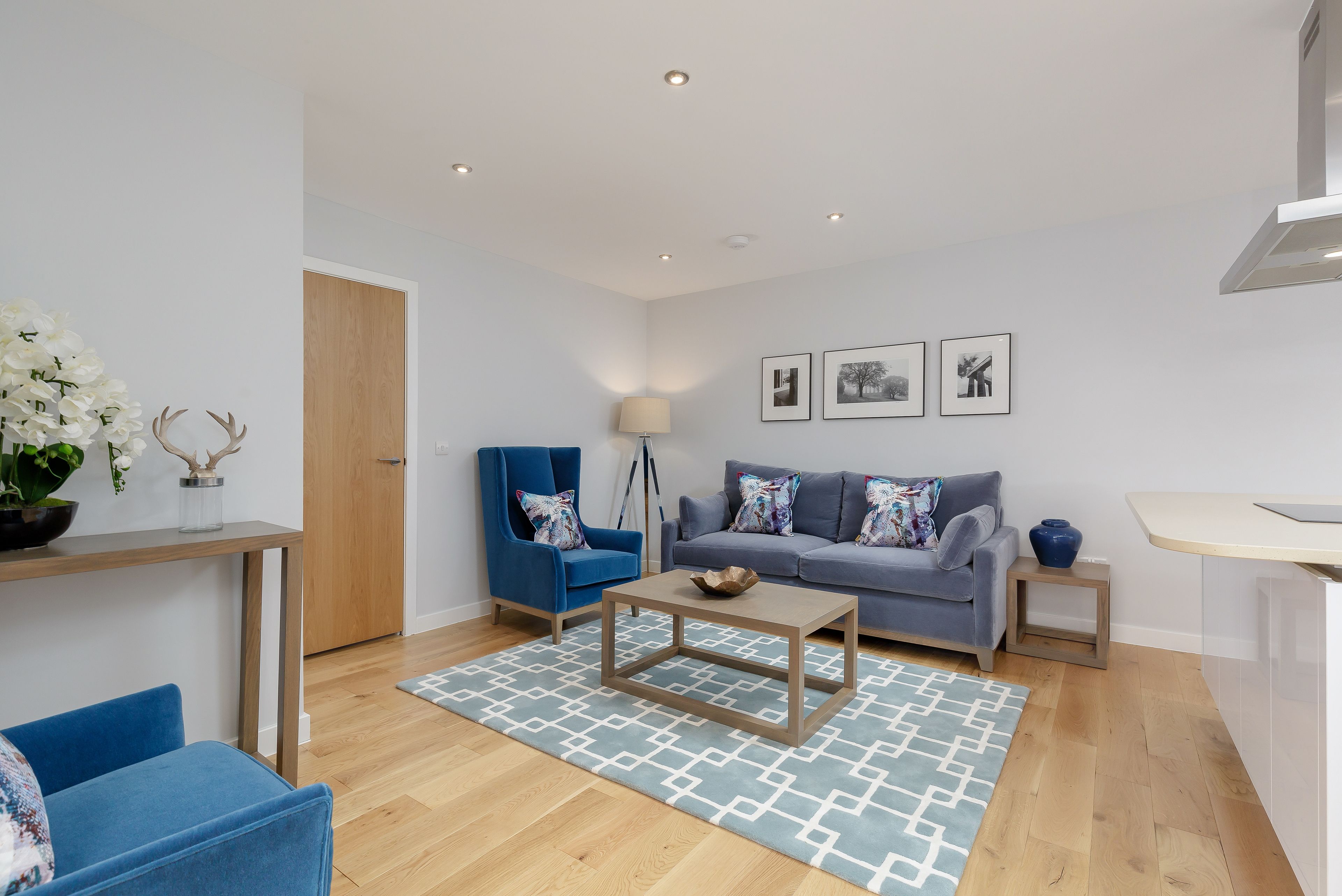 A modern living room with a grey sofa, blue armchair, and a coffee table. There is a wooden door, a small side table with a vase, and a blue rug.